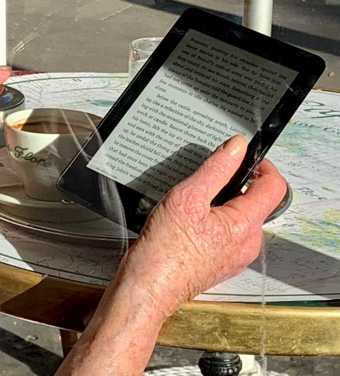 Bir kafe masasında okuyan bir el; yanında kahve fincanı, cam masanın üstünde şehrin yansıması.